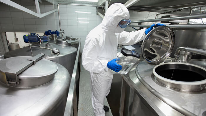 A biomanufacturing facility, a gowned technician about to pour liquid into a stainless steel sterile vat