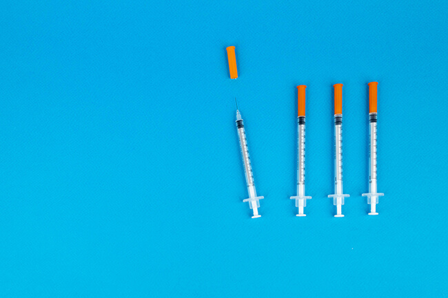 4 pre-filled syringes in a row against a blue surface. the 3 right syringes have orange caps and the left has the cap off just above the suringe.
