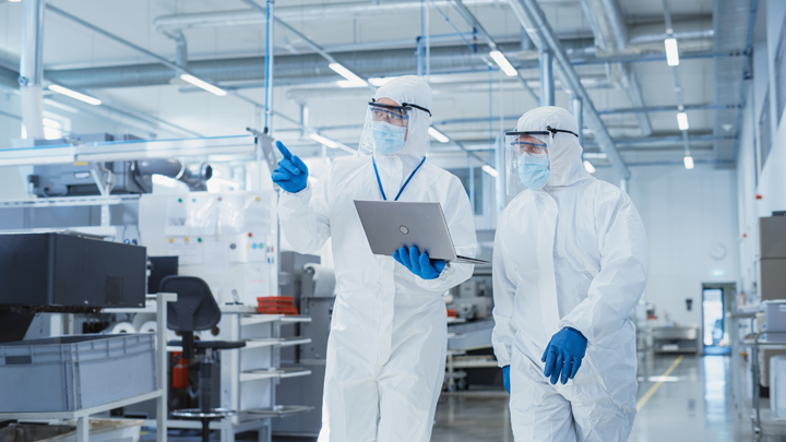 Two technicians in full bio gowns, gloves, masks and goggles walking a manufacturing floor, the one on the left holding a clipboard