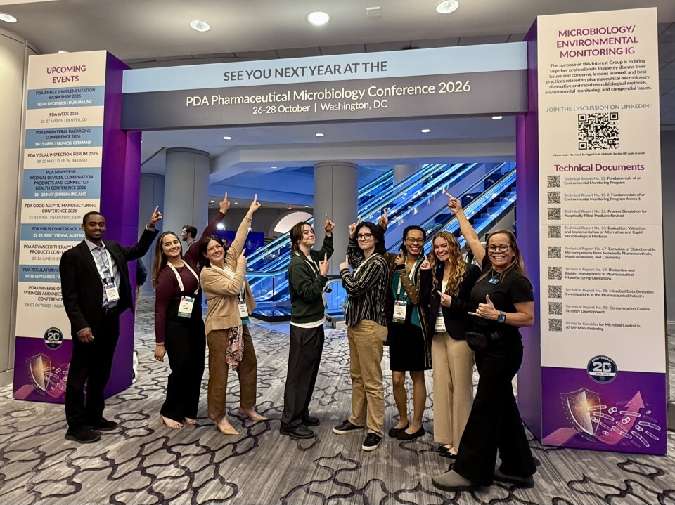 A group of student attendees at the 2025 Microbiology Conference standing and waving underneath the convention signage