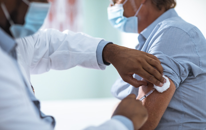 A physician in a white lab coat administering a vaccination in the upper left arm of a patient in a blue buttonup shirt