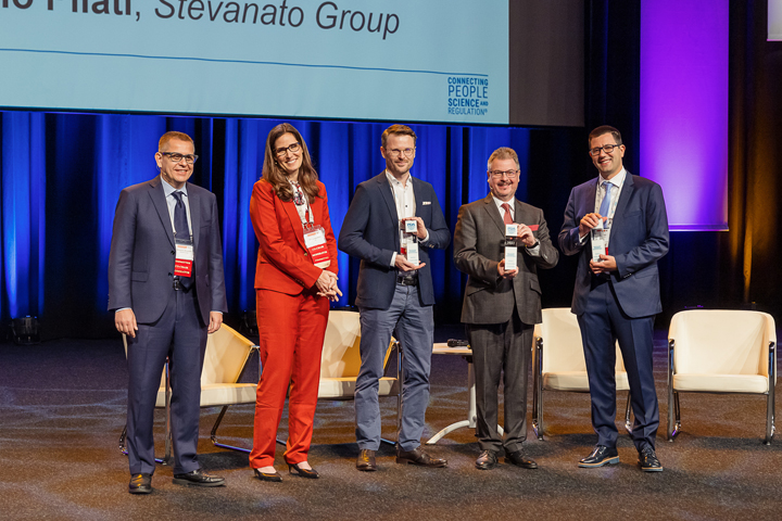 5 people standing on a stage, 3 men on the right holding up their awards