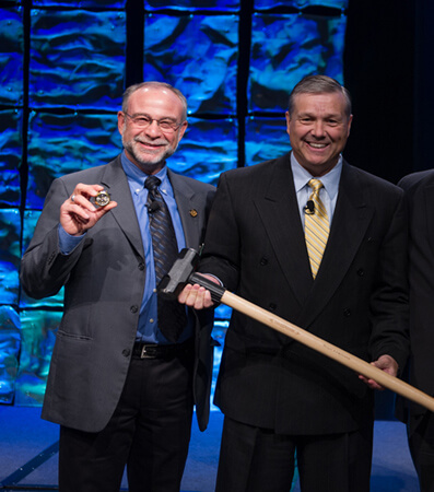 Hal Baseman on left holding the watch, Martin VanTrieste in center with a sledgehammer, speaker Marty Nealy on right